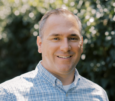 A man with short light brown hair, wearing a blue and white checked shirt, smiles outdoors with blurred green foliage in the background.
